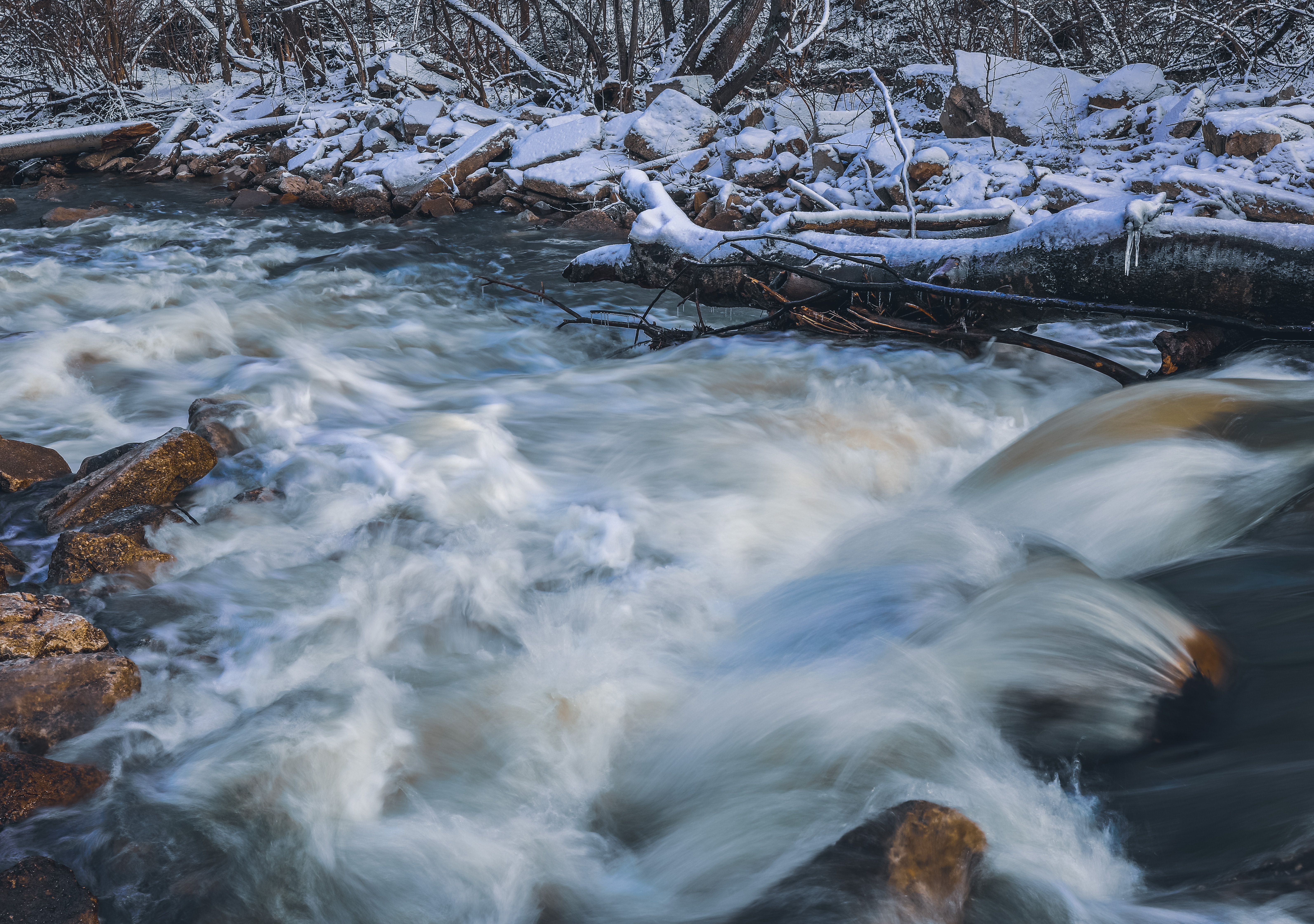Long-exposure winter cascades flowing past snow-covered rocks and fallen timber at Tawawa Park in Sidney Ohio
